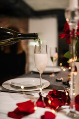 Champagne pours in glasses, flowers. Table setting for couple, Valentine's Day evening, burning candles for surprise marriage proposal. Candle light date in restaurant. Romantic dinner setup at night.