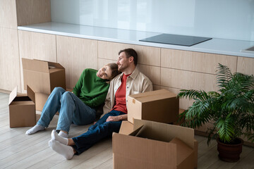 Happy tired couple sitting on floor in new own flat, house, resting, surrounded by cardboard boxes...