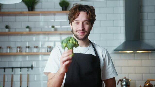 Smiling curly hair vlogger man chef in an apron recording video cooking lesson about vegetarian meal in modern kitchen. Showing vegetables one by one