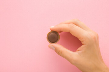 Young adult woman fingers holding brown caramel chocolate candy on light pink table background. Pastel color. Sweet snack. Closeup. Top down view.