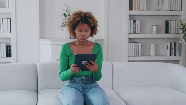 Young upset African American woman in casual clothes reading bad news on tablet computer and feeling stressed and depressed due to misinformation on internet sits on white sofa in apartment