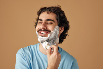 Portrait of young positive brunette man with curly hair applies shaving foam with retro shaving...