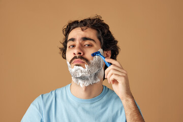 Portrait of young brunette man with curly hair applies shaving beard against sandy studio...