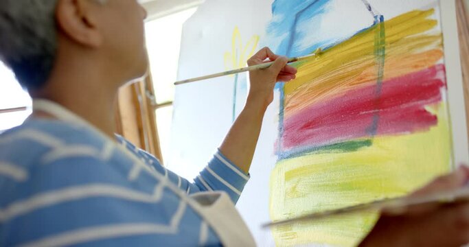 Focused senior biracial woman wearing apron and painting on big canvas at home, slow motion
