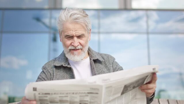 Positive aged male with beard enjoying reading newspaper in urban place while sitting on bench. Portrait of stylish senior person attentively searching article in morning press in good weather.