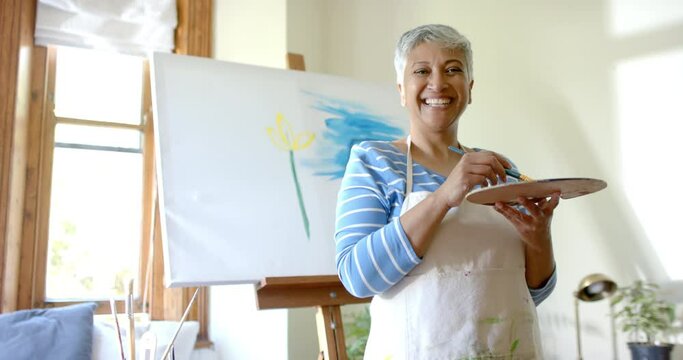 Portrait of happy senior biracial woman mixing colours on palette at home, slow motion