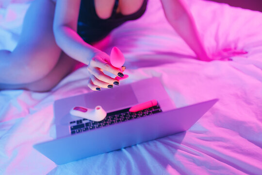 Beautiful woman lies in bed holding clitoral vibrator and watching porn on laptop, neon color. Women has Internet sex chat online