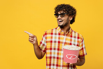 Young smiling happy cool Indian man wear 3d glasses watch movie film hold bucket of popcorn in cinema point index finger aside on area copy space isolated on plain yellow background studio portrait.