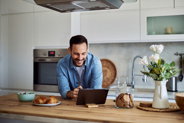 A smiling man reads on a tablet while leaning on a kitchen counter.