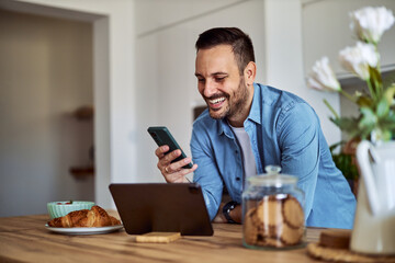 A happy freelance man uses a phone while leaning on a table in front of a tablet.