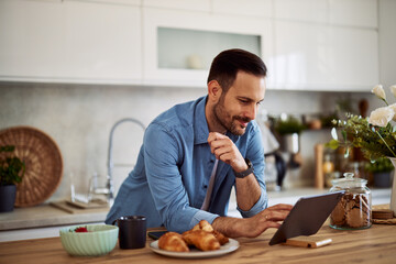 A focused adult man checking an email on a tablet while leaning on a kitchen counter.