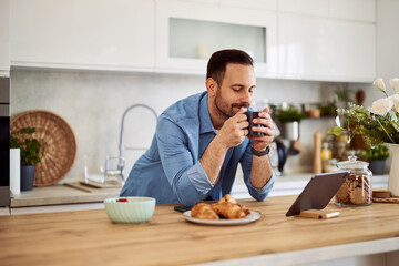 A content freelance eyes-closed man holding a cup of coffee in his hands while leaning on a kitchen counter in front a tablet.