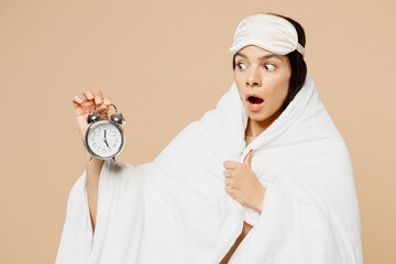 Young shocked Latin woman wrapped in blanket wears jam sleep eye mask rest relax at home hold in hand alarm clock isolated on plain pastel light beige background studio. Good mood night nap concept.