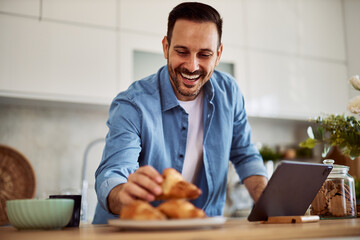 A happy freelance man leaning forward to take a croissant while standing in front of a tablet.