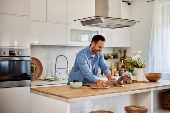 A happy male freelancer laughs and uses a tablet in a kitchen setting.
