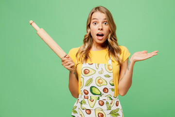 Young shocked housewife housekeeper chef cook baker woman wear apron yellow t-shirt hold rolling pin spreading hand look camera isolated on plain pastel green background studio. Cooking food concept.