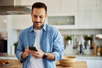 A content young adult man texting his friends on his phone in a kitchen setting.