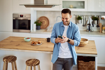 A smiling freelance man sending his coworker a funny text message while standing in a kitchen.