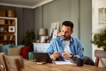 A focused male online virtual accountant looking at a tablet and holding a pen with a sheet of...