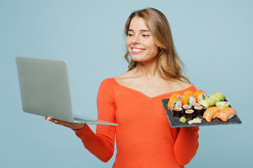 Young happy IT woman in orange casual clothes use work on laptop pc computer hold eat order raw fresh sushi roll served on black plate Japanese food isolated on plain blue background studio portrait