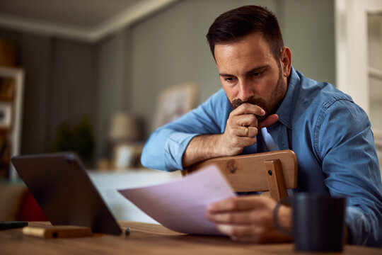 A focused young adult male freelancer analyzing the documents in his hand with a hand over his mouth.