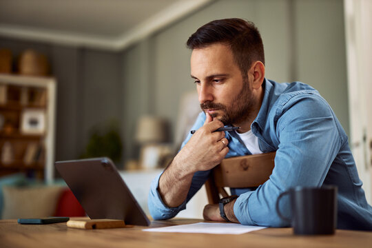 A deeply focused male freelancer looking at a tablet with a hand on his chin and sitting backwards in a chair.
