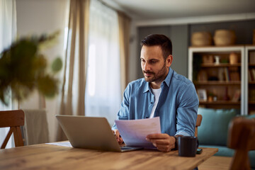 A focused freelance man making a video call with a client to discuss his latest project using his laptop and holding documents in his hands.