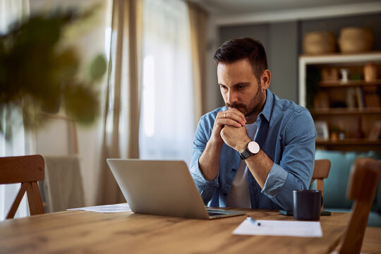 A deep-in-thought male freelance writer revising his latest work on a laptop with his hands connected on his chin.
