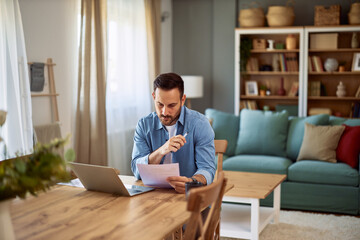 A focused male remote worker analyzing data on a paper he is holding while sitting at a desk in...
