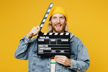 Young cheerful happy man he wear denim shirt hoody beanie hat casual clothes hold in hand classic black film making clapperboard isolated on plain yellow background studio portrait. Lifestyle concept - obrazy, fototapety, plakaty