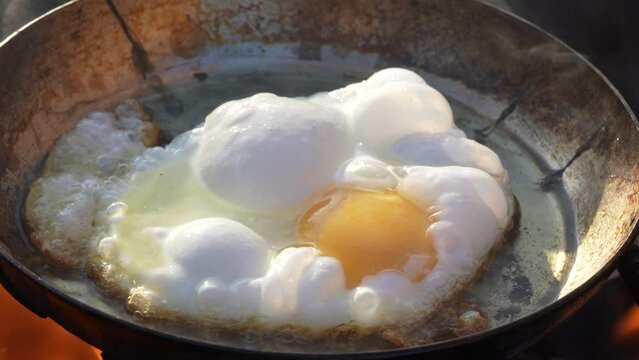 Fresh egg is fried in a frying pan outdoors with a sizzling oil and smoke on an open fire, close up