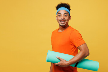 Side view fun young fitness trainer instructor sporty man sportsman wear orange t-shirt hold in hand yoga mat spend time in home gym isolated on plain yellow background. Workout sport fit abs concept.