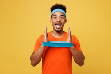 Young surprised fitness trainer instructor sporty man sportsman wear orange t-shirt use elastic band on hands spend time in home gym isolated on plain yellow background. Workout sport fit abs concept.