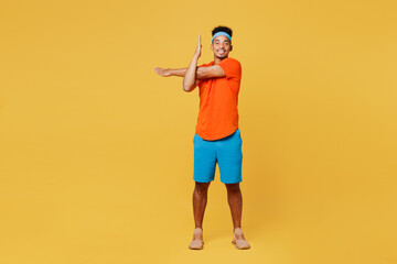 Fototapeta premium Full body happy young fitness trainer sporty man sportsman wear orange t-shirt do stretch exercise for hand spend time in home gym isolated on plain yellow background. Workout sport fit abs concept.