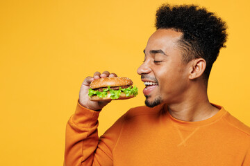 Close up young happy fun man of African American ethnicity wear orange sweatshirt casual clothes hold eat bite fast food burger isolated on plain yellow background studio portrait. Lifestyle concept.
