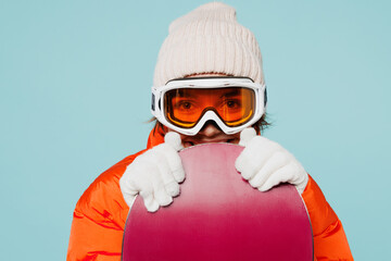 Close up young fun woman wear warm padded windbreaker jacket hat ski goggles mask hold snowboard looking camera travel rest spend weekend winter season in mountains isolated on plain blue background - obrazy, fototapety, plakaty