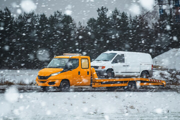 A rollback tow truck transports a broken car during a snowstorm. Car failure in winter. A tow truck on the public road.