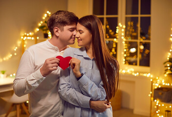Tender loving couple enjoying Valentine's Day. Happy man and woman spend time together, give each other presents and red heart shape paper cards, kiss, relax, dance to slow music in room with lights