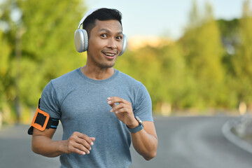 Fototapeta premium Happy millennial man listening to music in headphones, jogging in the park on a sunny day