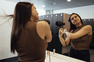 Smiling brunette woman using hairdryer at sport club after training