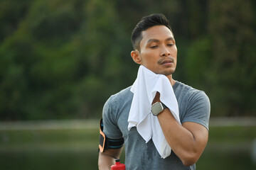 Young muscular man resting and wiping sweat with a towel after morning cardio workout in park - obrazy, fototapety, plakaty