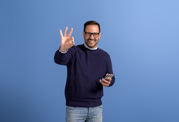 Fototapeta premium Portrait of smiling salesman using mobile phone and gesturing OK sign on isolated blue background