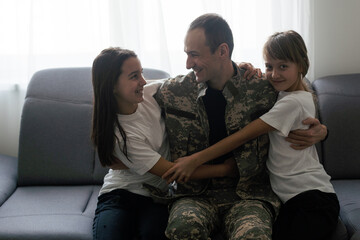  happy positive smiling soldier man in camouflage sitting with his daughter on sofa, looking how...
