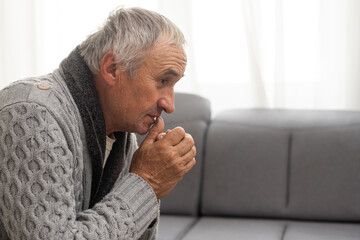 Senior man with white hair looking very sad while sitting on the armchair and thinking about something in his room