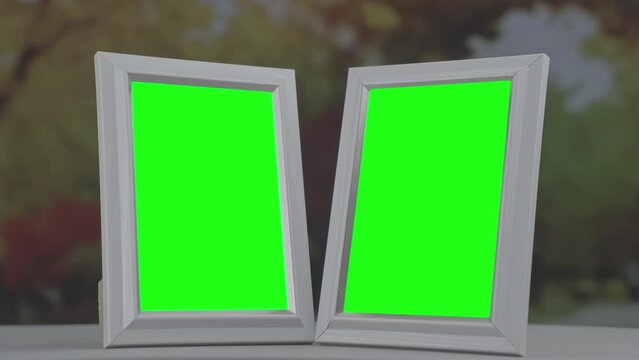 Pair of wooden photo frames with green chroma key space placed at home table in autumn season with view on falling golden leaves behind window glass.
