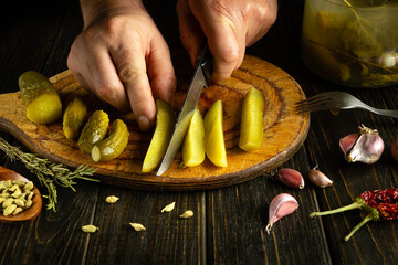 A chef's hand uses a knife to cut a pickled cucumber on a wooden cutting board. Working environment...