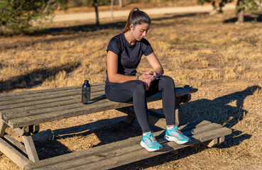 Naklejka na ściany i meble Young runner girl in sportswear sitting on a wooden bench in a park, enjoying listening to music with white wireless headphones looking at her results on her smart watch.