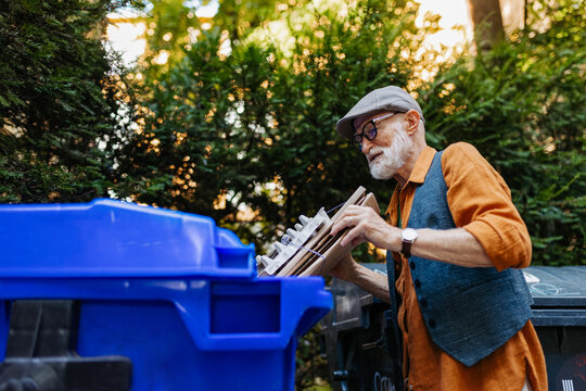 Senior man throwing paper waste, cardboards into recycling container in front apartment. Elderly man sorting the waste according to material into colored bins.