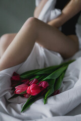 A beautiful young sexy girl in black underwear lies in bed in the morning with a beautiful spring bouquet of pink-red tulips, a holiday gift for a woman