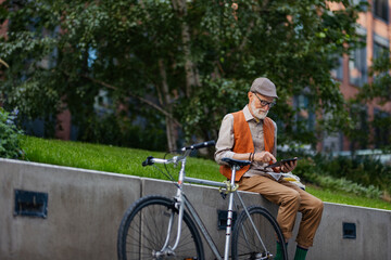 Fototapeta na wymiar Stylish senior man sitting in the city park, holding tablet, shopping online. Concept of digital literacy and seniors using digital technologies.